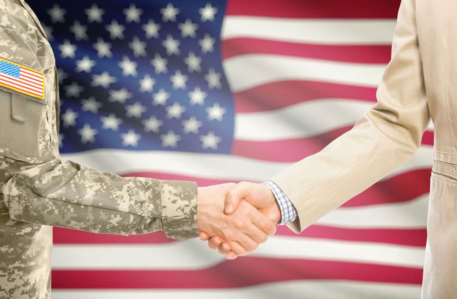 member of the military shaking hands with a civilian with American Flag in the background